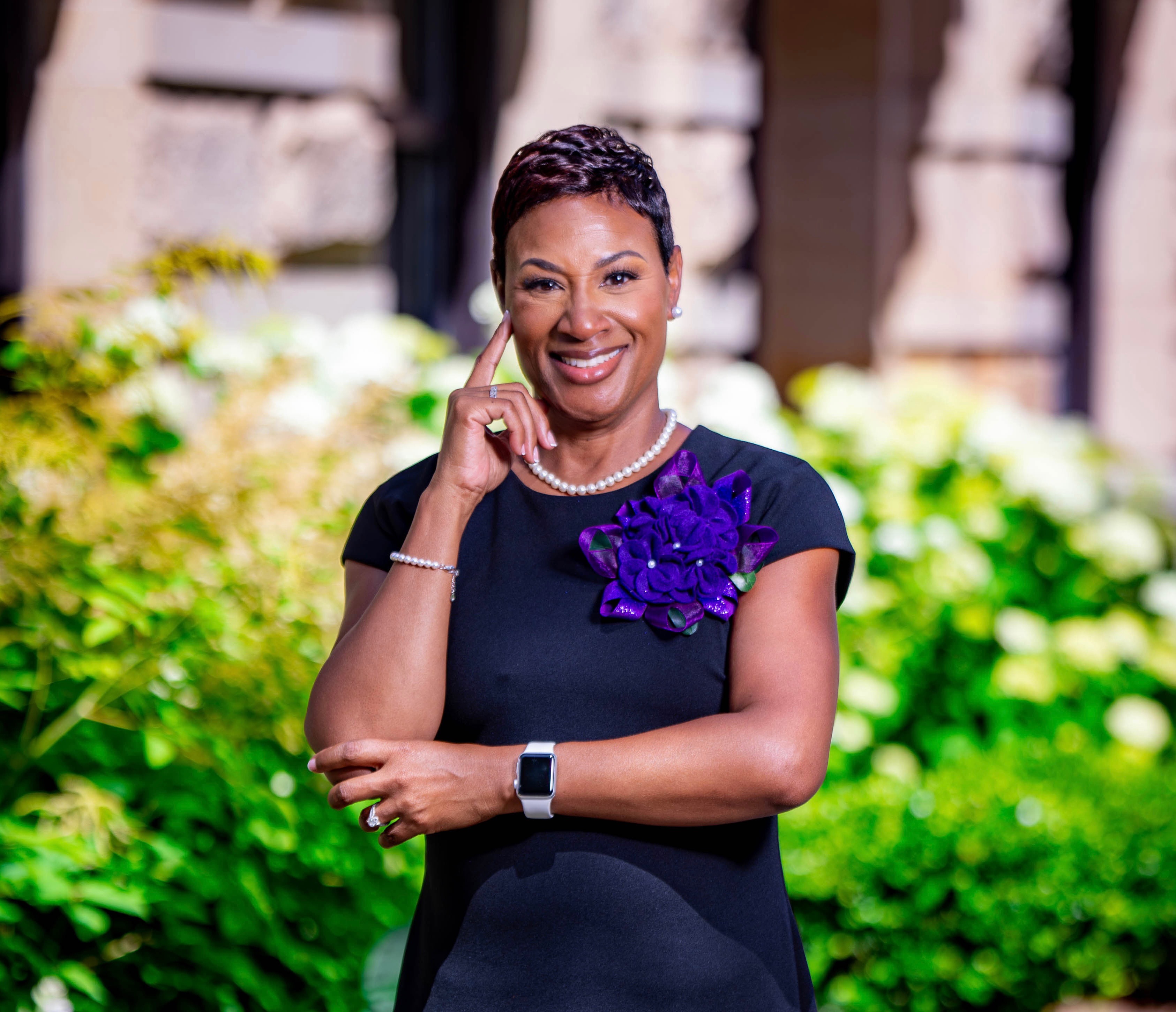 Black woman, with short pixie cut, in a black dress, wearing a purple violet corsage, standing outside